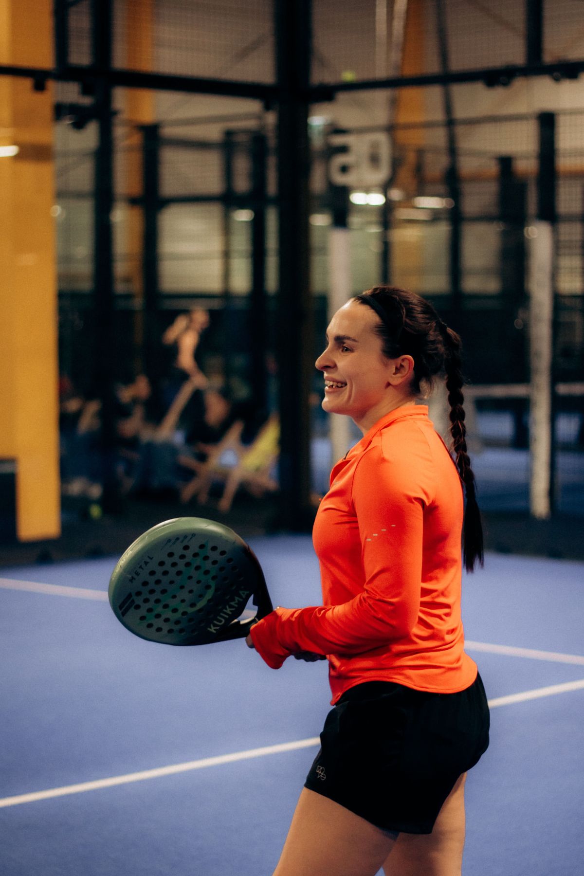A padel player in an orange long-sleeve top smiles as she turns with her paddle on a blue indoor padel court.