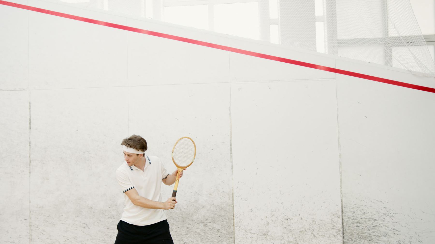 A player in a white polo and headband follows through on a shot against the white plaster wall of a squash court, with the red out-of-court line running along the side wall.